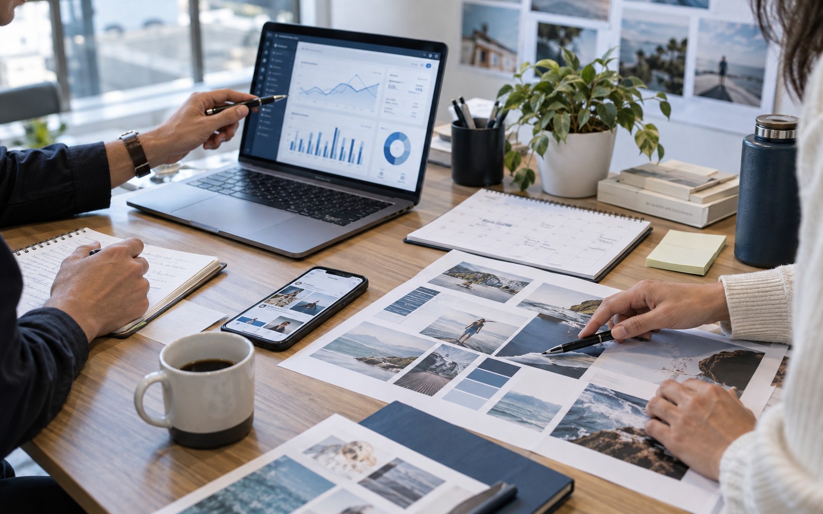 A real small business marketing planning workspace, laptop displaying a campaign performance dashboard with simple charts, smartphone with coordinated social media creative, printed brand campaign layouts and a calendar on desk, two colleagues' hands collaborating.